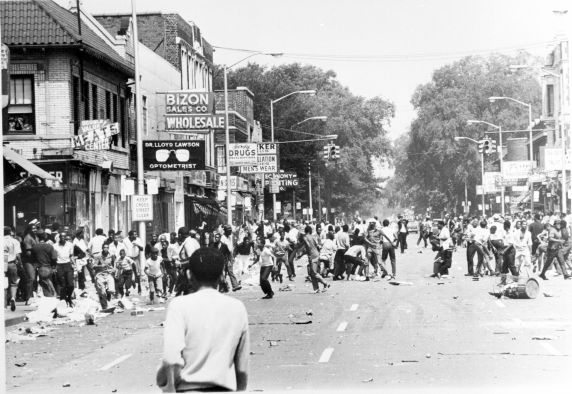 12th Street descends into chaos on the first day of the civil unrest of 1967. At rear, center, the Blind Pig, whose raid by police officers sparked the conflict, can be seen behind the Economy Printing sign. In the distance a photographer snaps pictures of the scene.